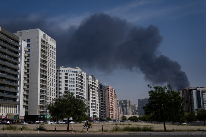 سقوط آرامش دبی؛ چگونه جنگ ایران تصویر لوکس خلیج فارس را بر هم زد؟ A cyclist rides past buildings in Sharjah City with a large plume of black smoke rising in the background.