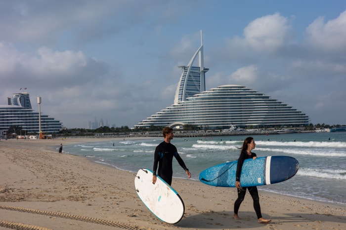 سقوط آرامش دبی؛ چگونه جنگ ایران تصویر لوکس خلیج فارس را بر هم زد؟ Two people in wetsuits carry surfboards on Jumeirah Beach with the Burj al Arab hotel in the background.